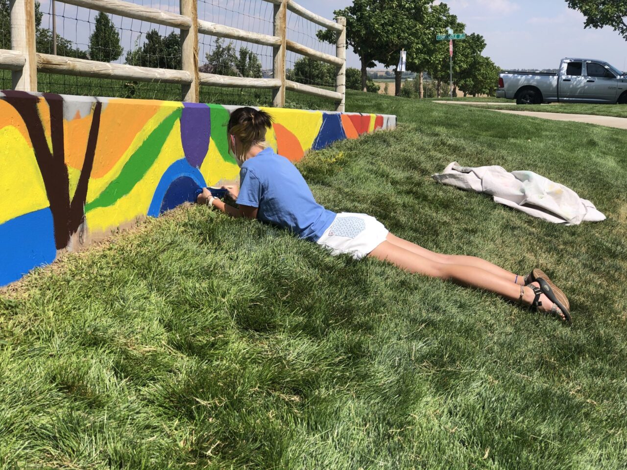 A woman painting a concrete barrier mural. She is lying flat on her stomach and applying the color blue.