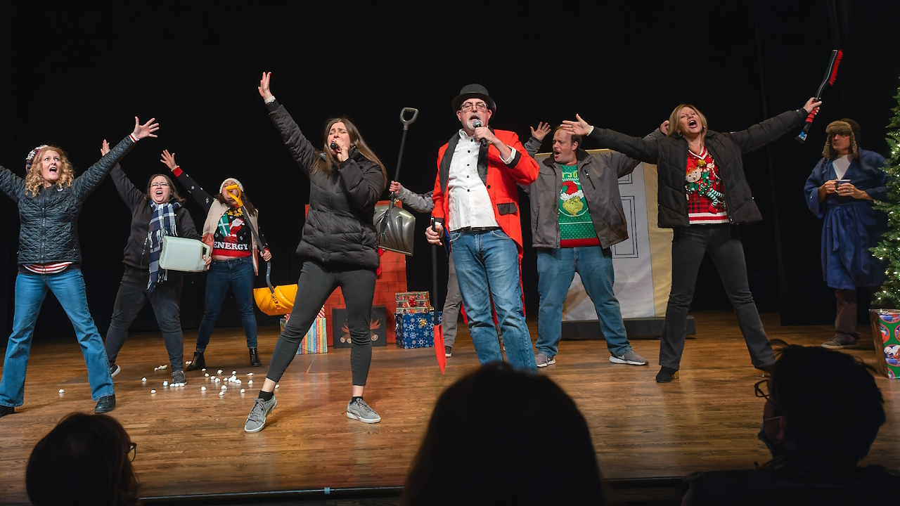 Actors singing and dancing on stage in Christmas sweaters while holding snow removal equipment